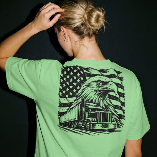 A woman viewed from behind wearing a light green t-shirt with a large black graphic on the back featuring an eagle, semi-truck, and American flag. She has blonde hair in a bun and is posing against a dark background.