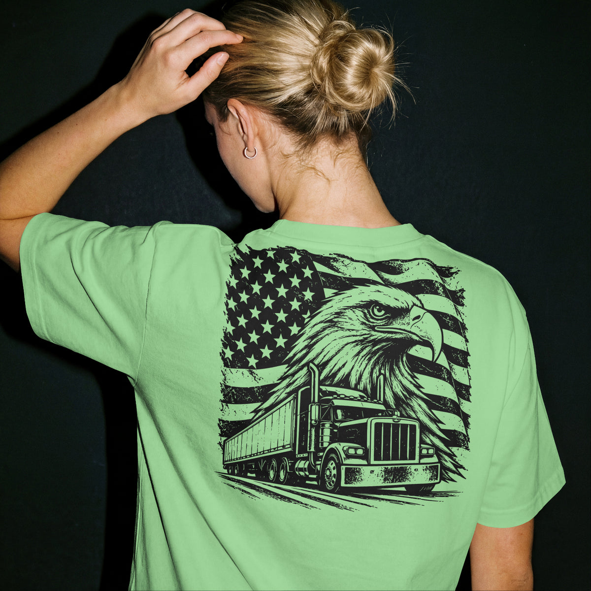 A woman viewed from behind wearing a light green t-shirt with a large black graphic on the back featuring an eagle, semi-truck, and American flag. She has blonde hair in a bun and is posing against a dark background.
