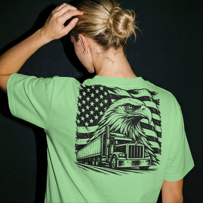 A woman viewed from behind wearing a light green t-shirt with a large black graphic on the back featuring an eagle, semi-truck, and American flag. She has blonde hair in a bun and is posing against a dark background.