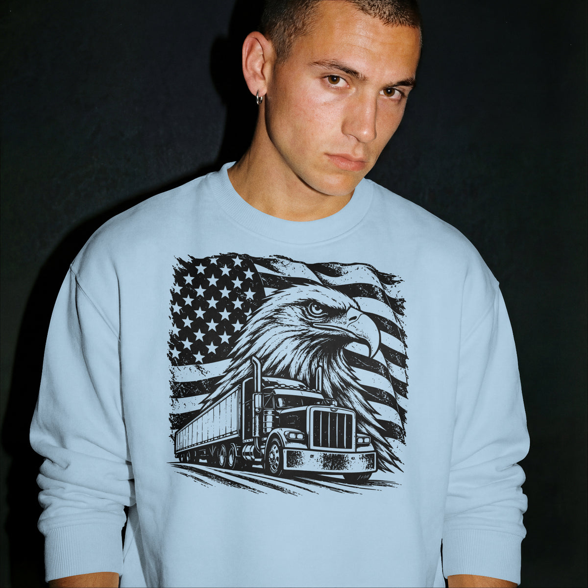 A man wearing a light blue crewneck sweatshirt with a large black graphic print of an eagle, American flag, and semi-truck on the front, set against a dark background.
