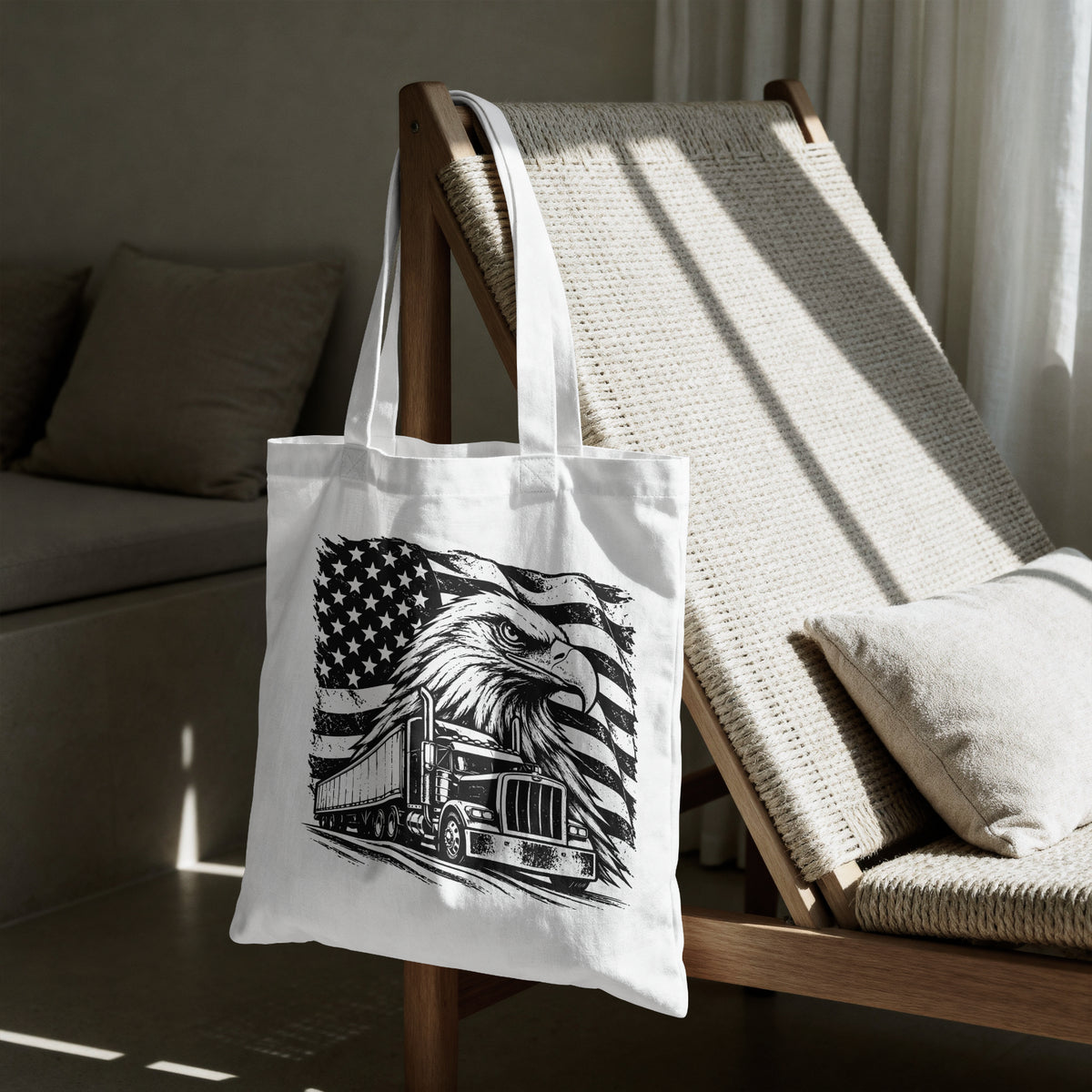 A white canvas tote bag with a black graphic print of an eagle and semi-truck over an American flag, hanging on a wooden chair in a sunlit living room.