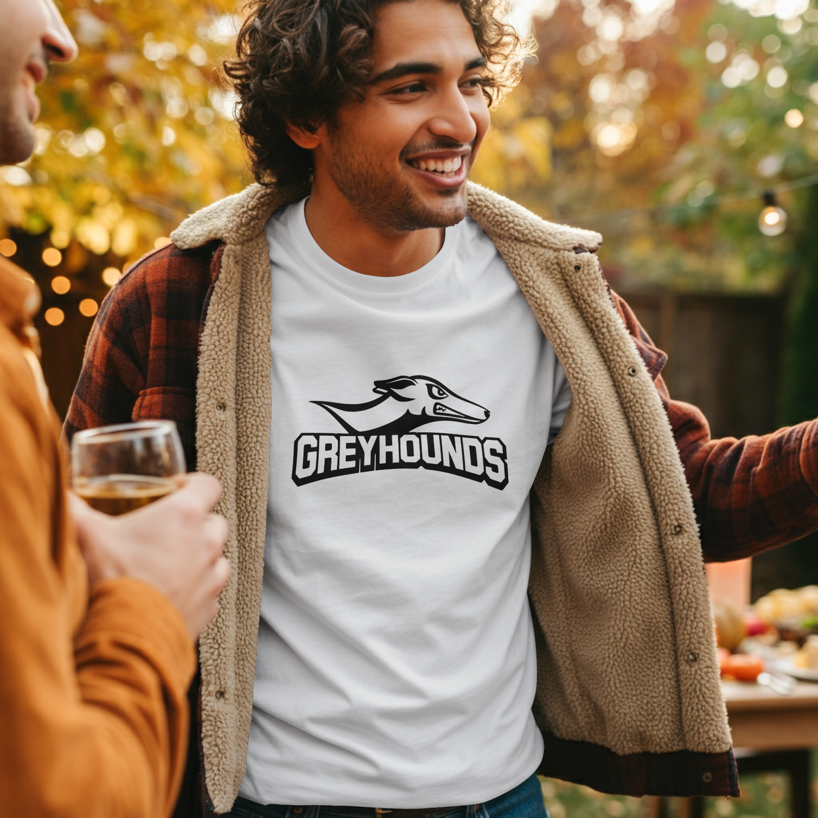 A man outdoors wearing a white graphic T-shirt with a black greyhound dog logo and the word 'GREYHOUNDS' across the chest, layered under a tan sherpa-lined flannel overshirt. Another person holding a drink is partially visible on the left, and the background shows a softly blurred outdoor setting with warm bokeh lights.