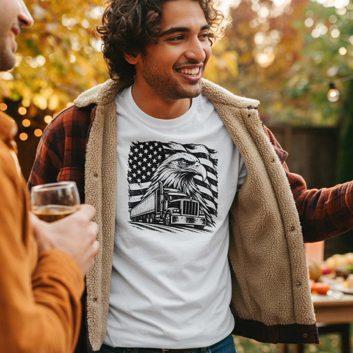 A man outdoors wearing a white graphic T-shirt with a black truck and American flag design, layered under an open tan fleece flannel overshirt and a red plaid flannel shirt. Another person holding a drink is partially visible on the left.