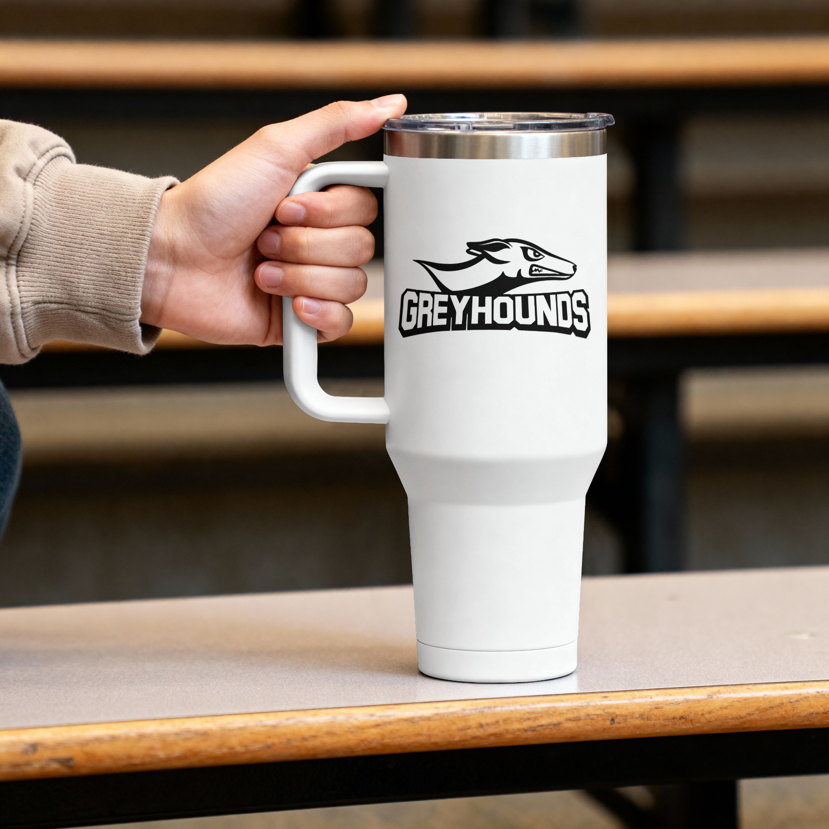 A white insulated travel mug with a stainless steel lid and handle, featuring a black 'GREYHOUNDS' logo with a hound head graphic. A hand wearing a beige sweatshirt is holding the mug, which is set on a wooden table in front of blurred wooden bleachers.