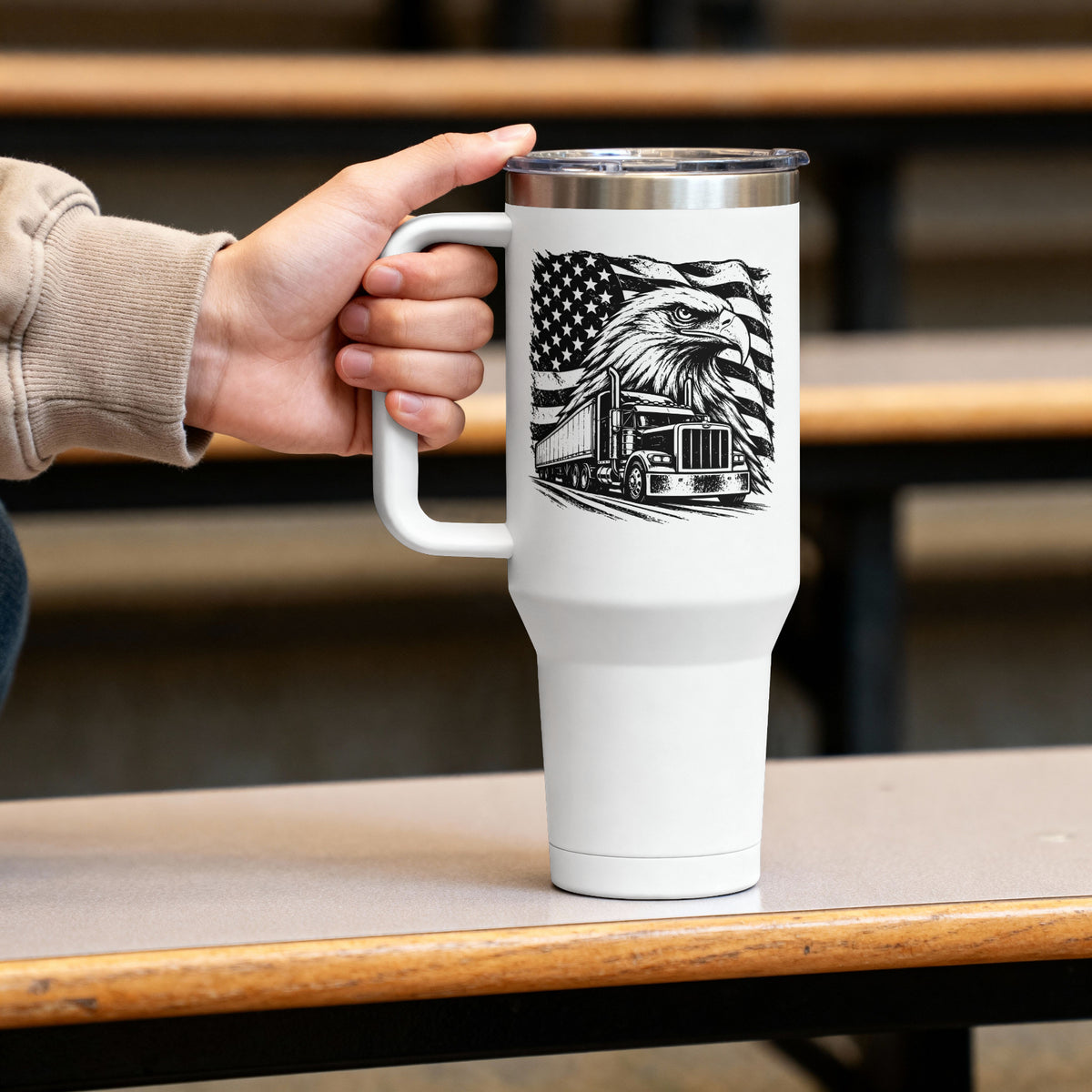 A white insulated travel mug with a handle and clear lid sits on a wooden table, held by a person's hand. The mug features a black graphic of an eagle head over a semi-truck with an American flag background.
