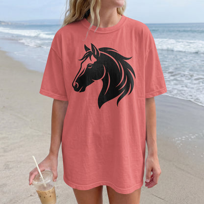 A woman wearing an oversized coral pink t-shirt with a black horse head graphic, standing on a sandy beach with the ocean in the background.