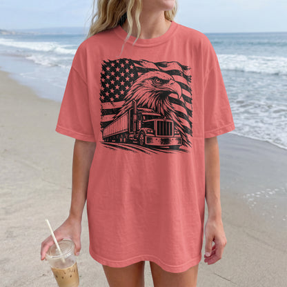 A woman standing on a beach wearing an oversized coral pink t-shirt with a black graphic of an eagle head, a semi-truck, and an American flag. She is holding an iced coffee.
