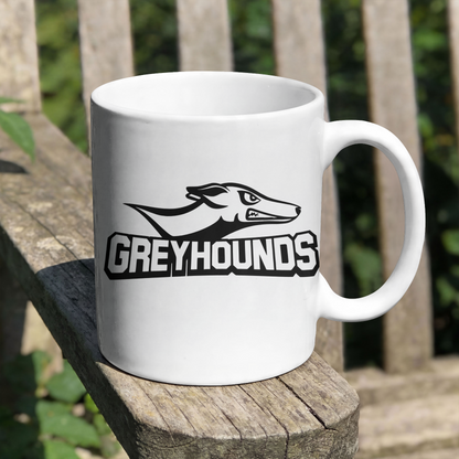 A white ceramic mug with a black and white greyhound graphic and the word 'GREYHOUNDS' printed on the side, resting on a weathered wooden bench with a blurred outdoor background.