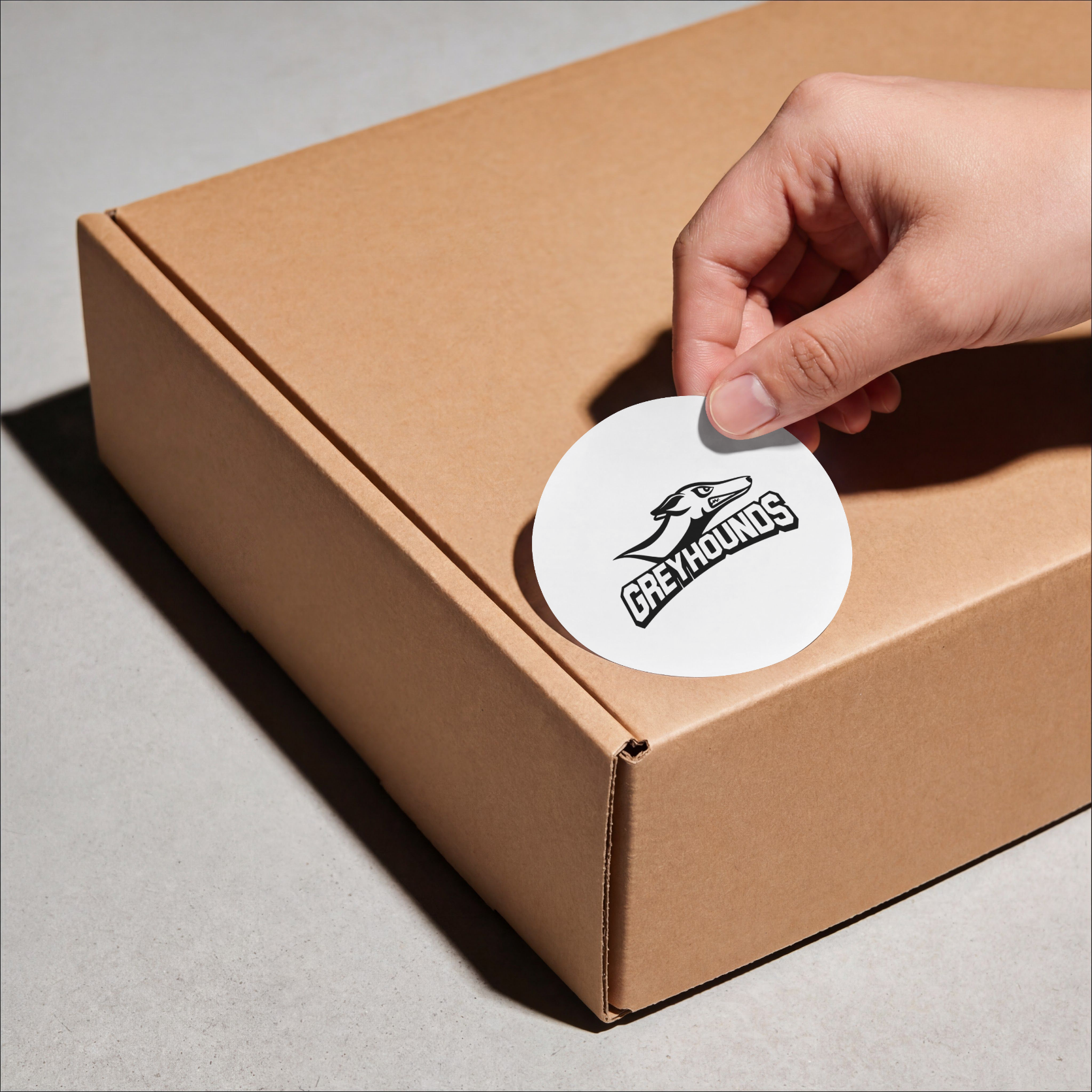 A hand is placing a round white sticker with a black greyhound logo and the word 'GREYHOUNDUS' onto a brown cardboard box. The sticker is branded as a Greysounds decal.